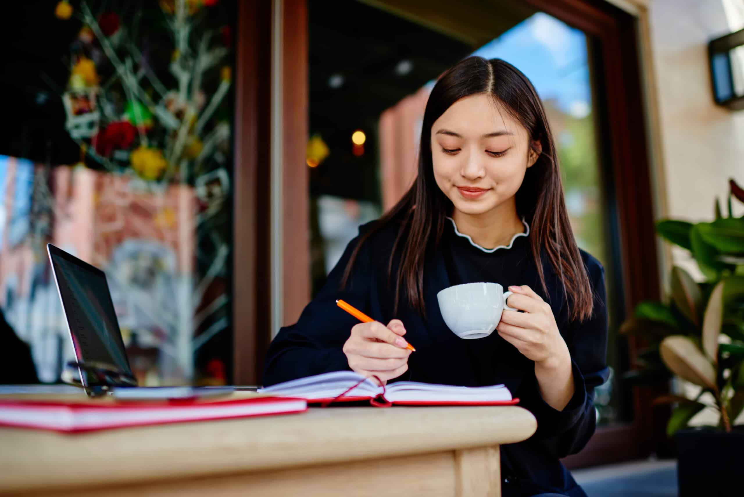 Asian woman writing in her notebook while drinking coffee.