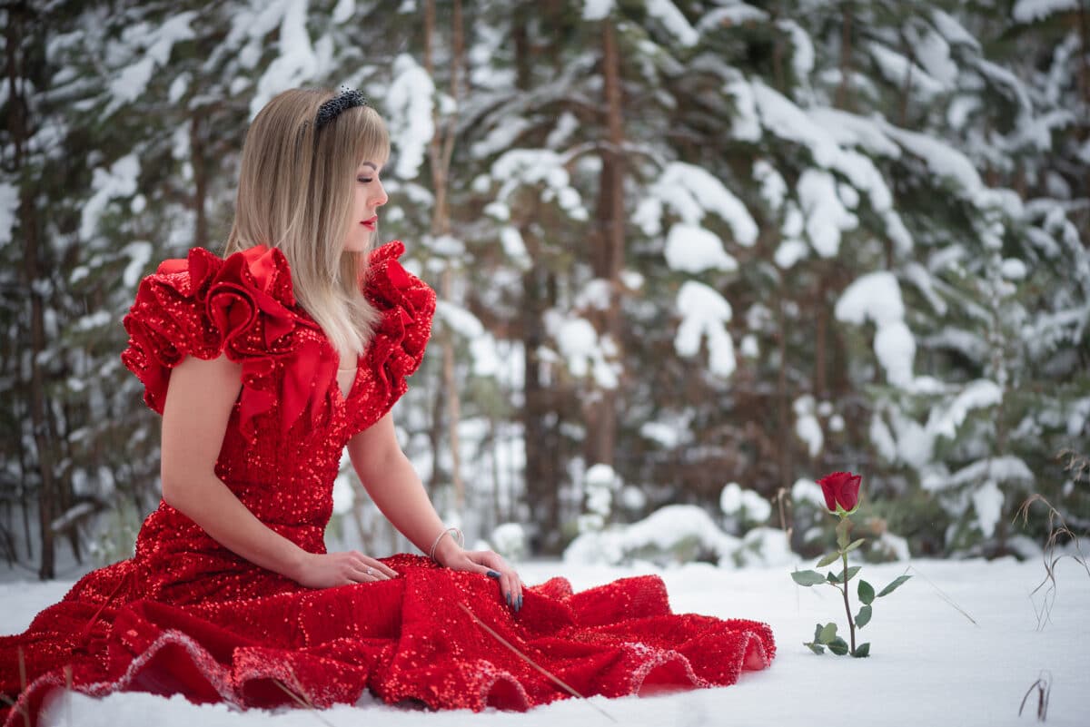 Young beautiful woman in the red dress with the red rose flower in the winter snowy forest. Winter fairytale concept.