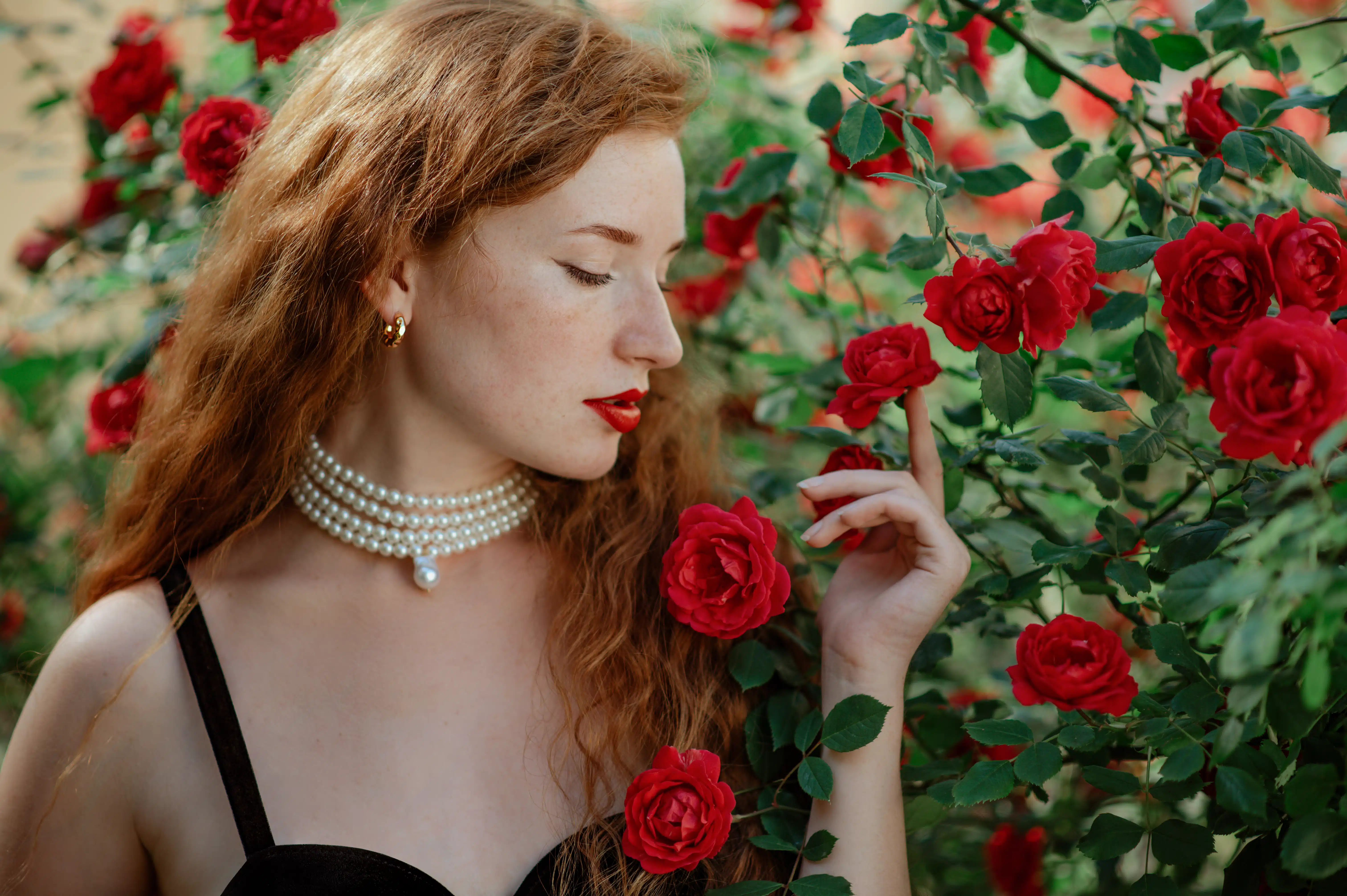 Beautiful redhead wearing elegant pearl necklace enjoying the roses in the garden