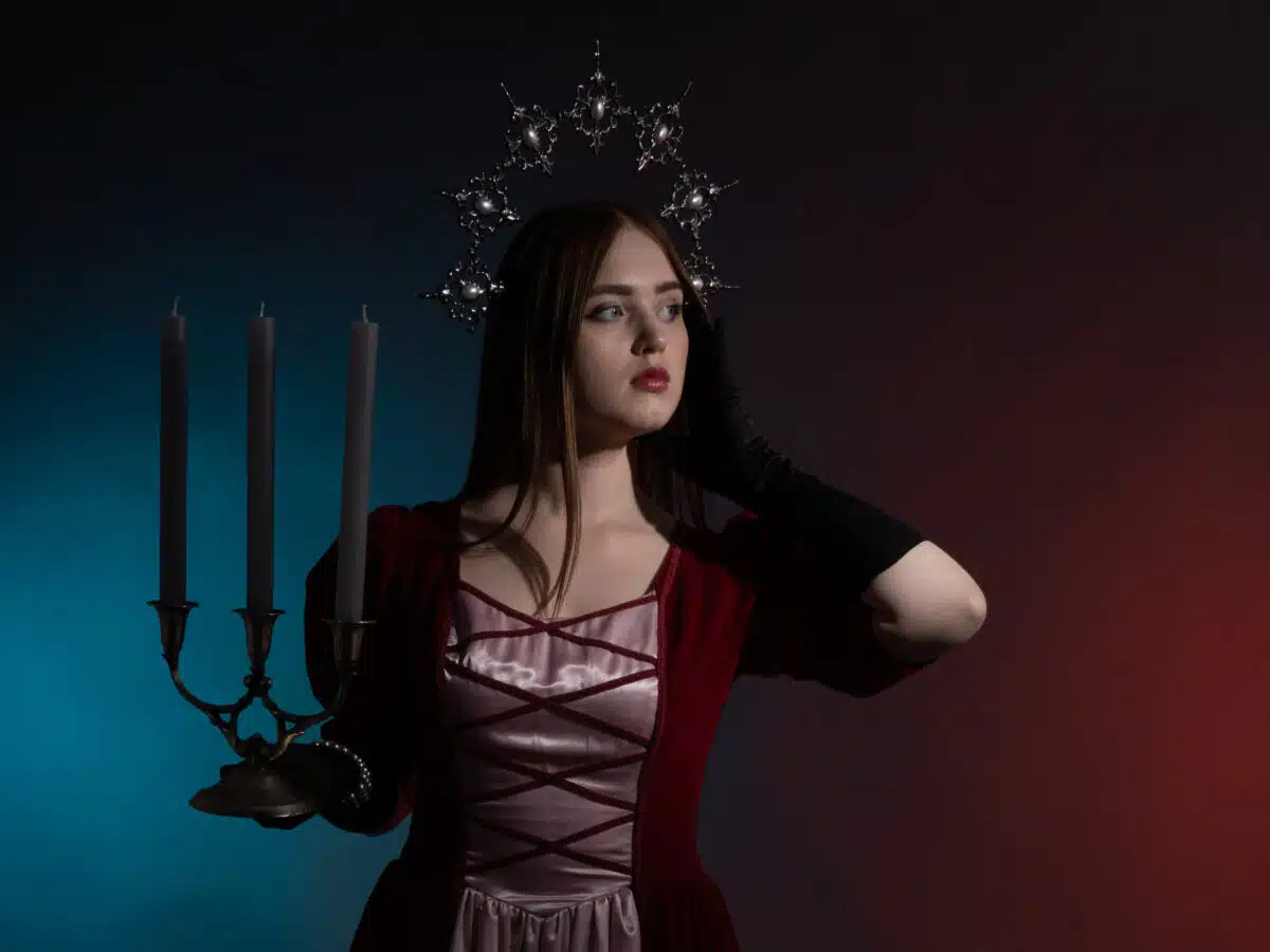 Mythical fantasy queen with a candlestick. Silvery gothic crown on the head. Burgundy vintage artistic dress, black gloves. Medieval style. Medium shot portrait in a dark key