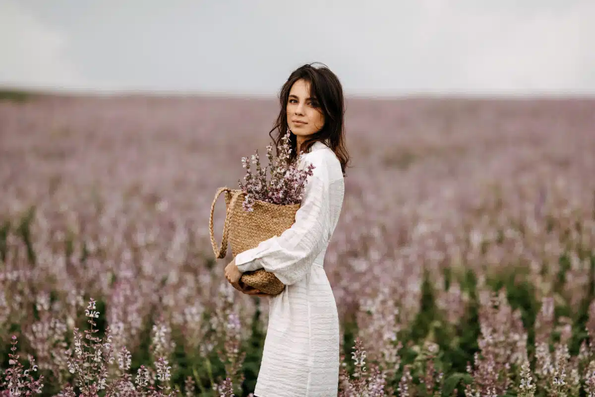 a young woman in white summer dress, standing in a big field with sage in bloom, holding a wicker bag with flowers