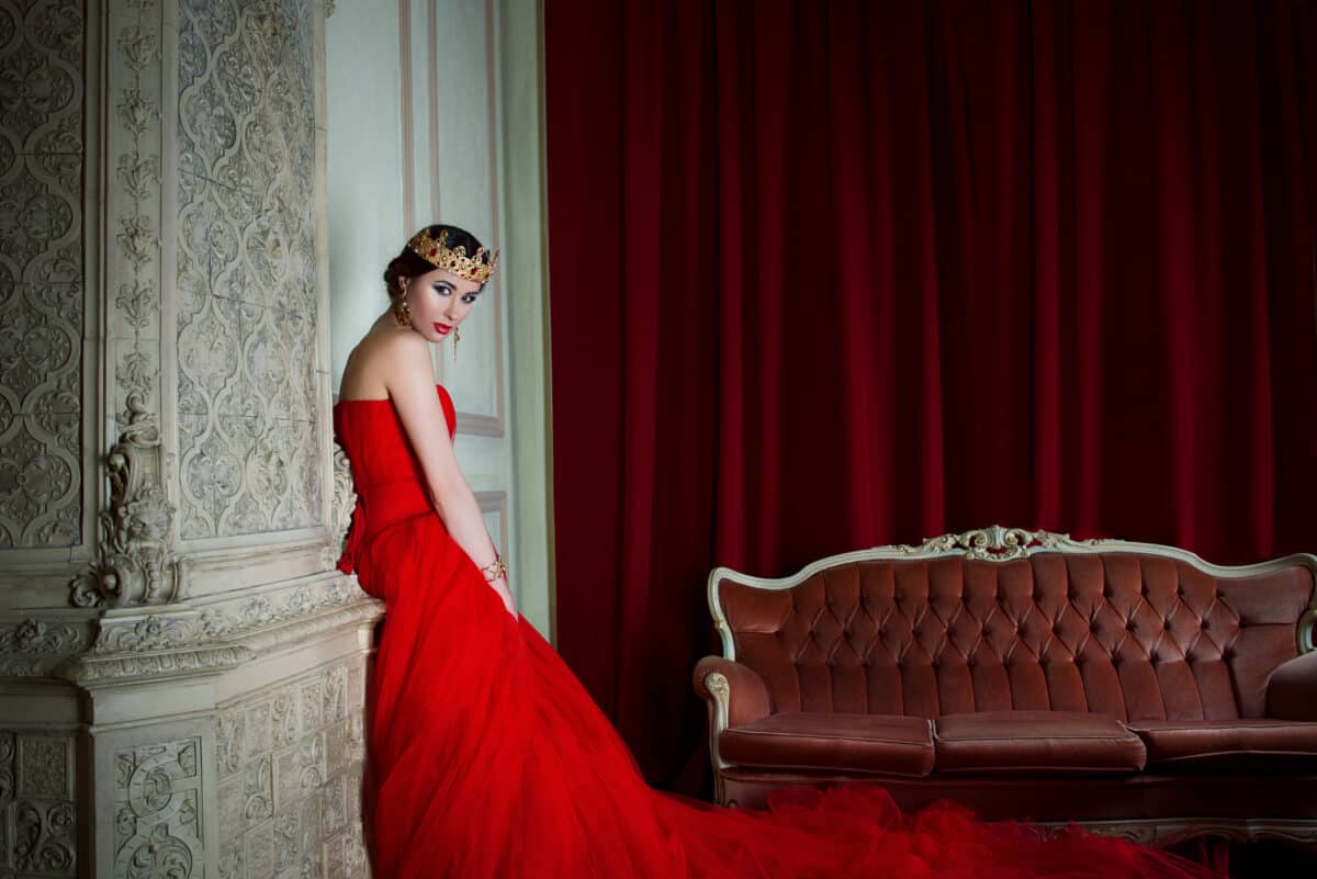 Beautiful queen in long red dress and royal crown stands near the fireplace