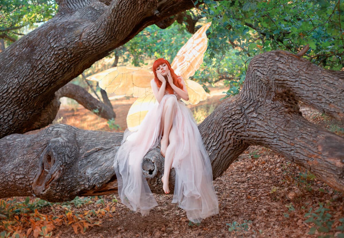 Young beautiful woman fantasy elf sits on branch of tree in spring forest. Green trees summer park. Pixie fairy girl. pink silk dress gold shiny glowing butterfly wings. Costume magic creation angel.