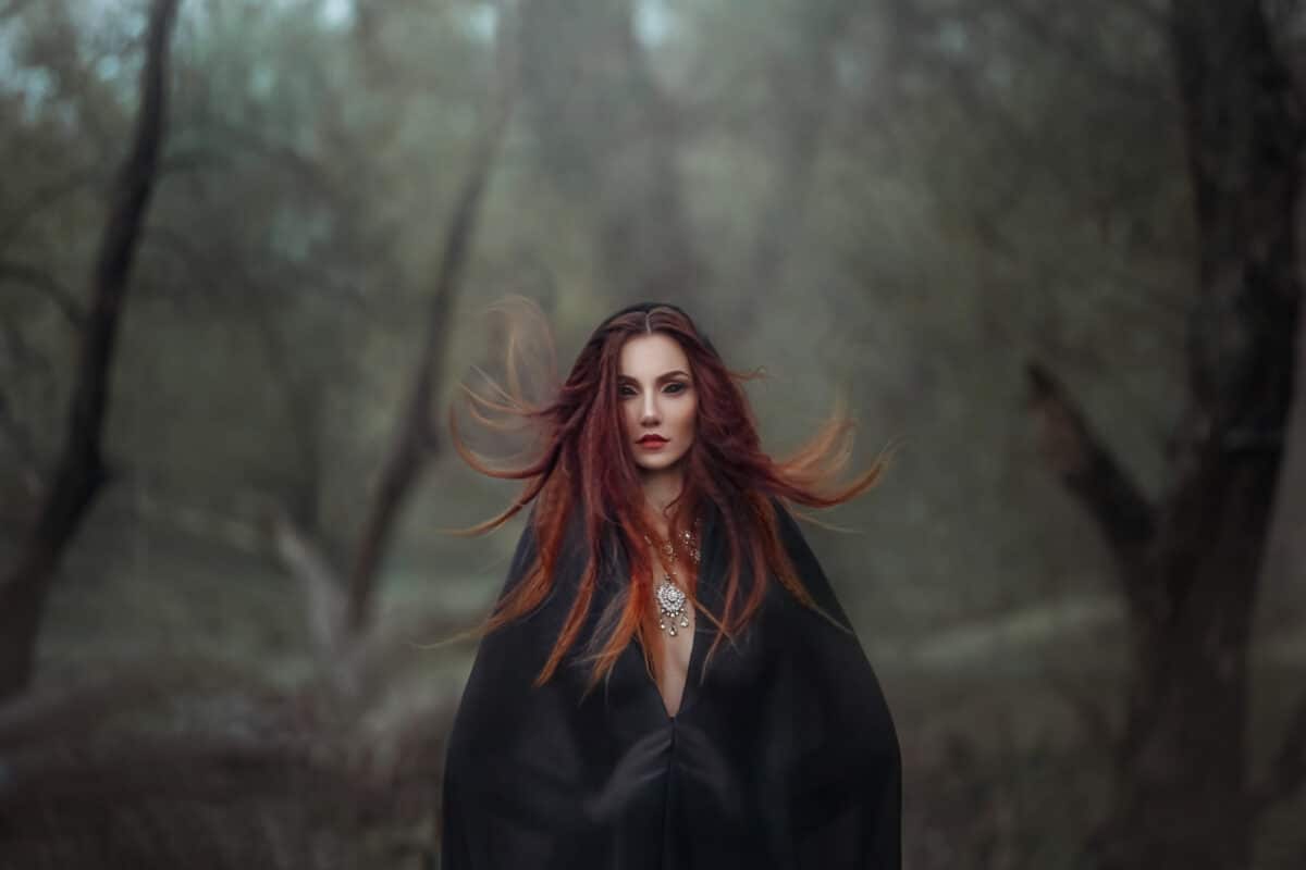 Mysterious fantasy gothic woman dark witch obsessed by evil. Red-haired Girl demon in black dress cape hood. Red hair flutters in wind. Dark dense deep forest background, trees. Scleral lenses on eyes.