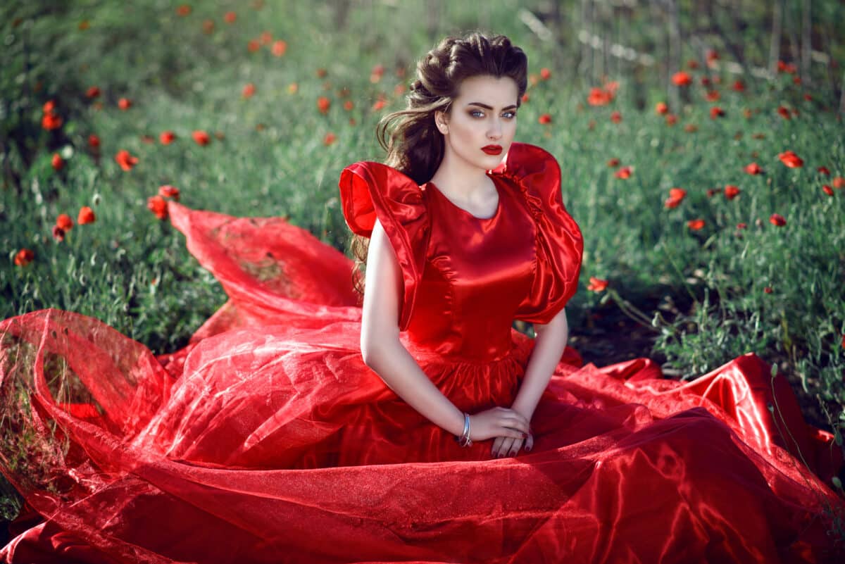 Beautiful blue-eyed young lady with perfect make up and hairstyle wearing luxurious silk red ball gown with ruffle sleeves sitting in the poppy field.