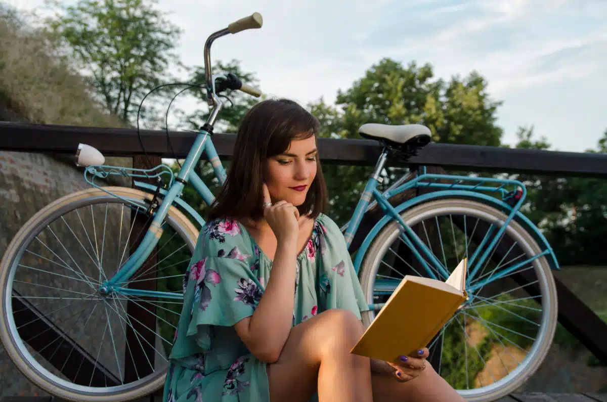 Cute woman in summer dress reading book outdoors next to a bicycle.