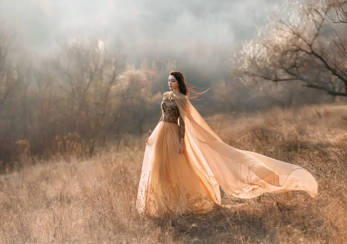 Pretty brunette girl walks in the forest . She is dressed in luxurious, golden dress. The wind playing with her hair and fabrics. The beautiful weather of autumn fog and sun.