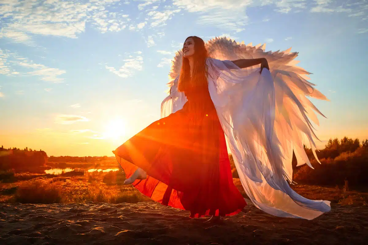 Beautiful young woman or girl in red dress and white wings on the sand on sunny day with blue sky. Angel model or dancer posing in photo shoot on dunes