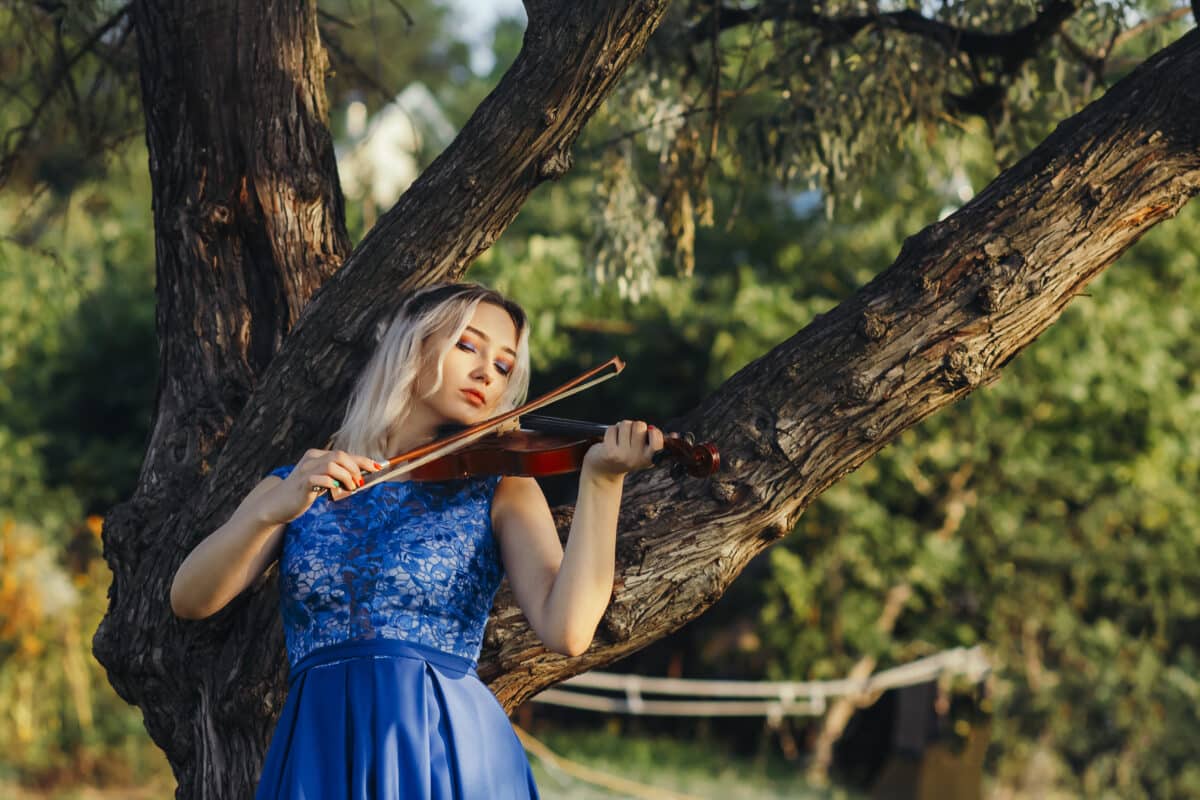 beautiful romantic girl playing violin lean on tree outdoors, young woman in long elegant dress with a musical instrument on nature, concept music and art