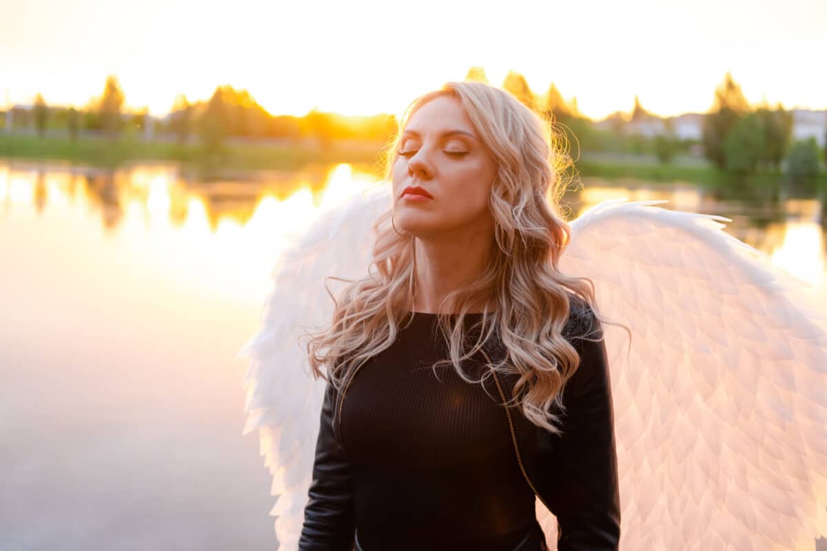 sexy blonde woman in black leather jacket and shorts with white angel wings. demon or angel in hell or heaven. sunset near lake