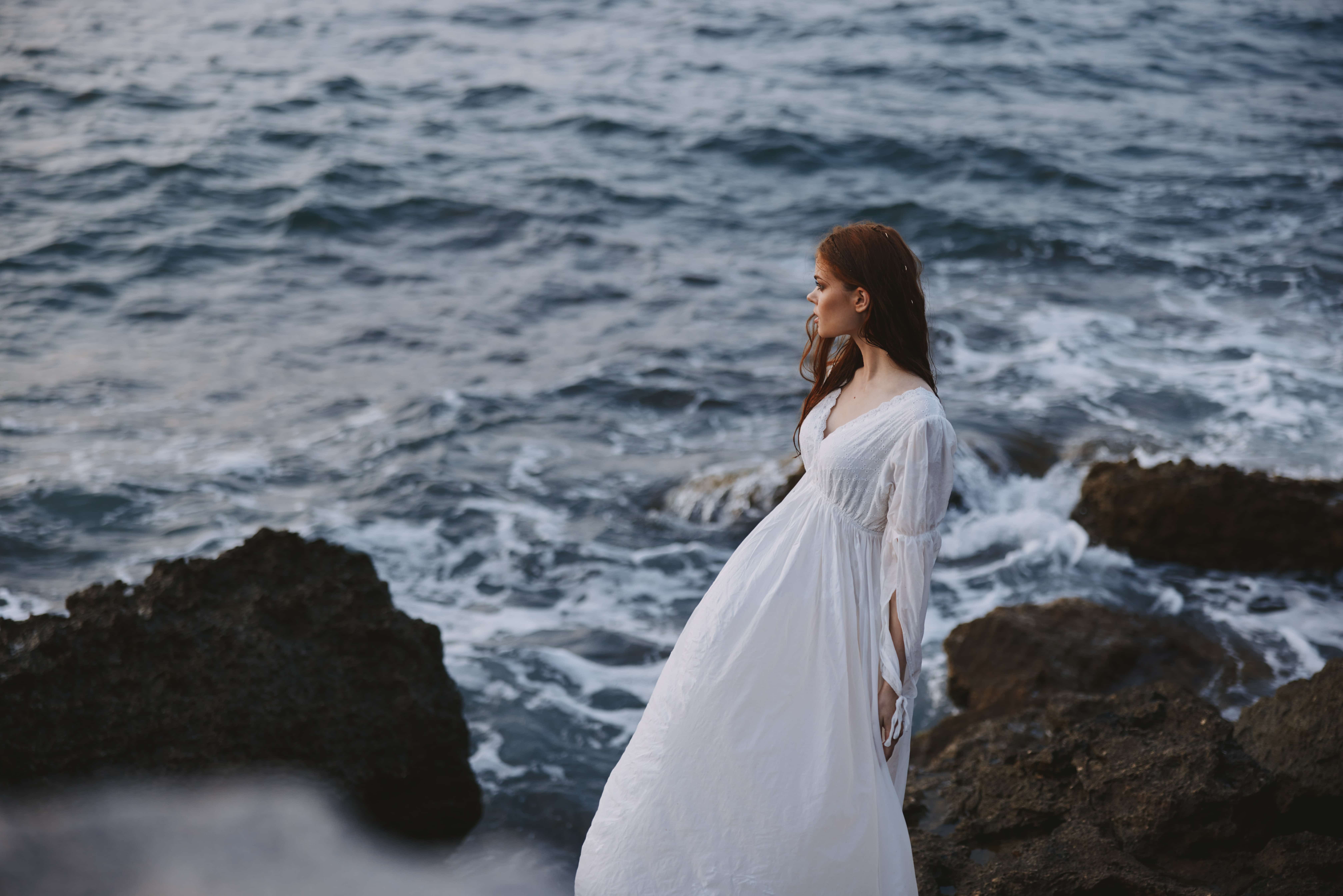 A woman in a white dress stands on the shore of the ocean stones 