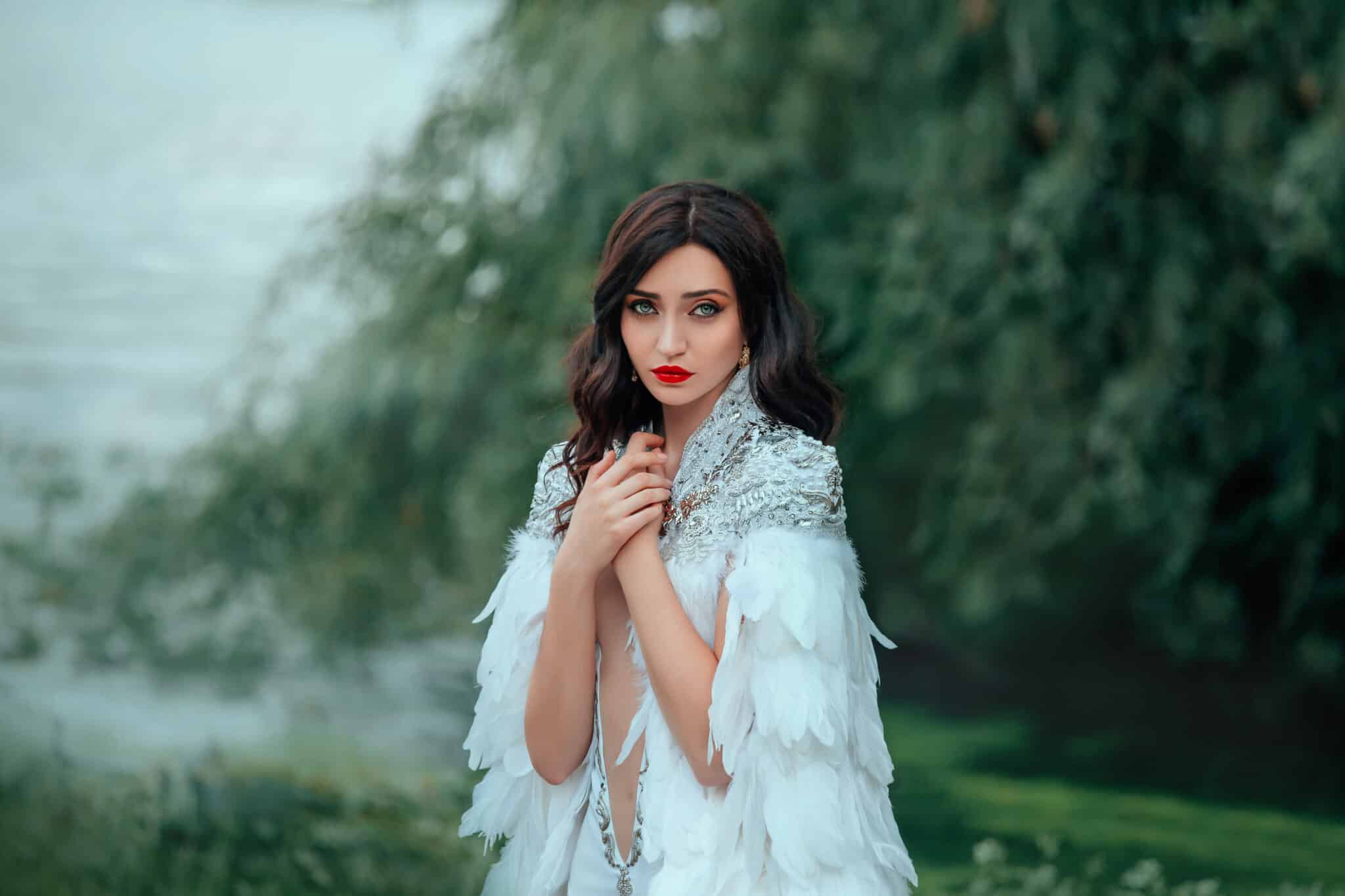 Portrait strong attractive brunette woman in luxury royal fantasy outfit. Make up red lips. long Cloak feathers white sexy dress deep neckline. Spring Haze over water river. Summer Green nature lake.