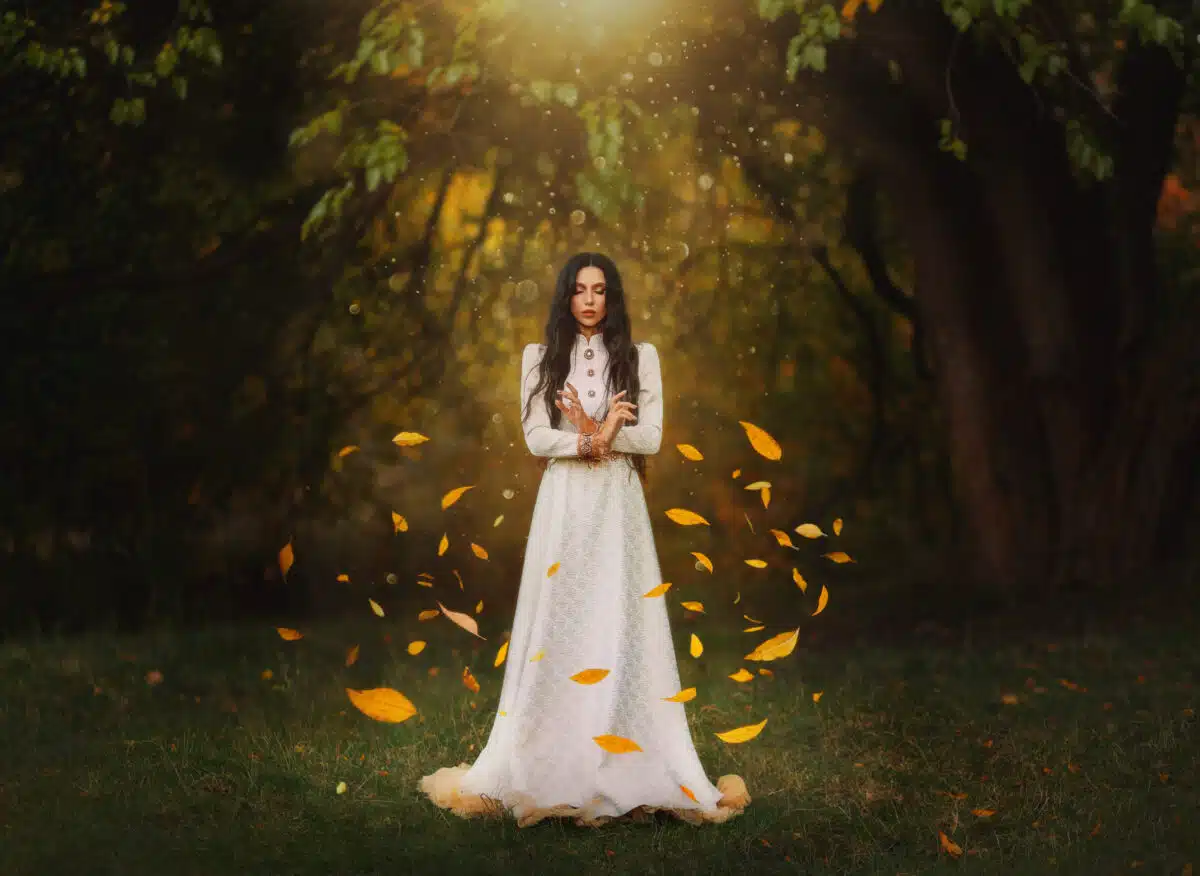 Gothic art photography fantasy woman creates magic. Bright divine light, autumn yellow leaves soar rotate fly in wind around fairy girl princess. Lady witch. white long dress. Dark forest black trees