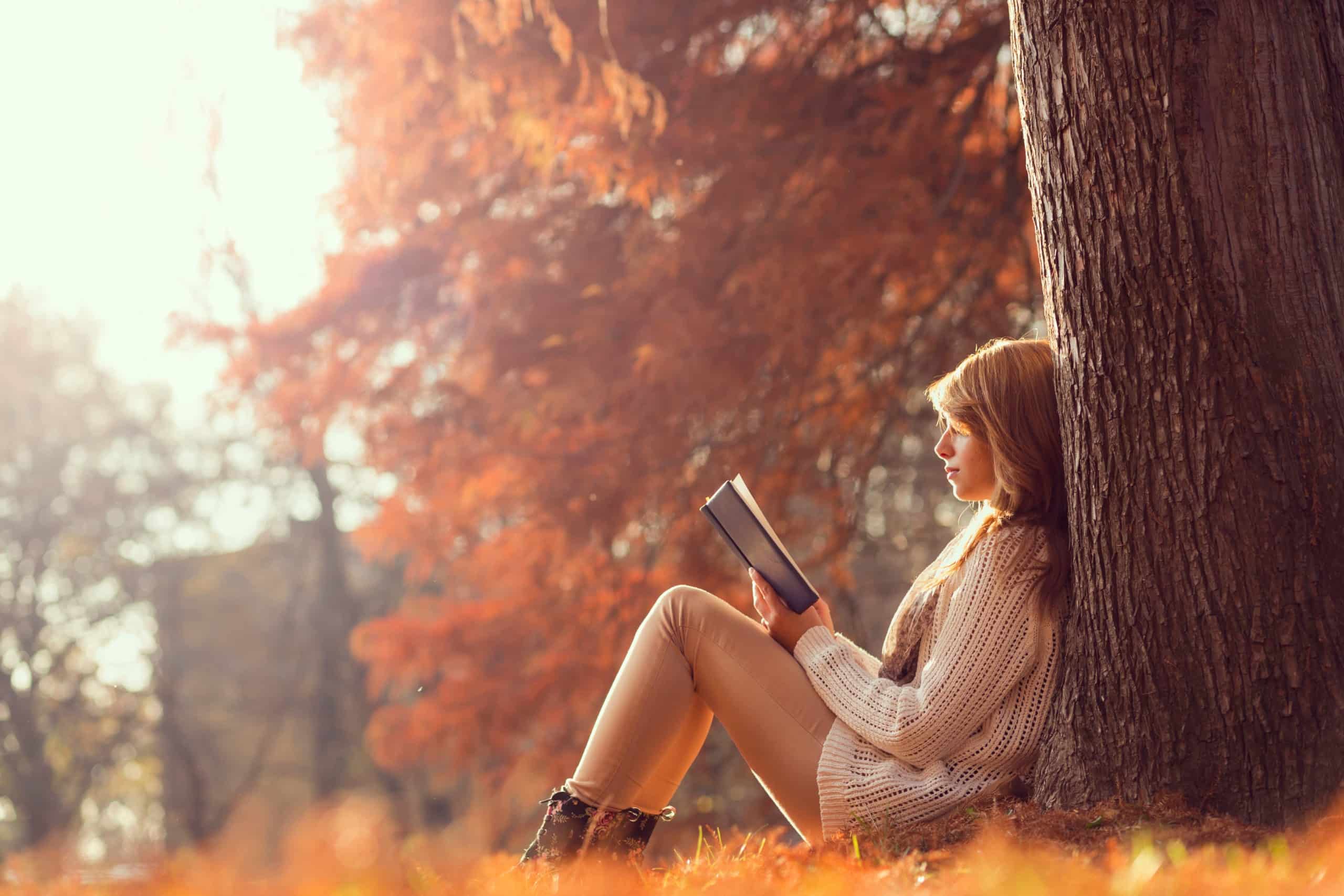 Young woman sitting on the grass, leaning against a tree, reading a book in autumn.
