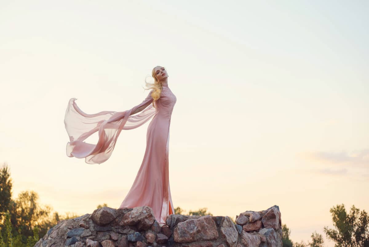 elegant princess with blond fair wavy hair with tiara on it, wearing a long light pink rose fluttering dress, standing on the tower of the castle at the sunset.
