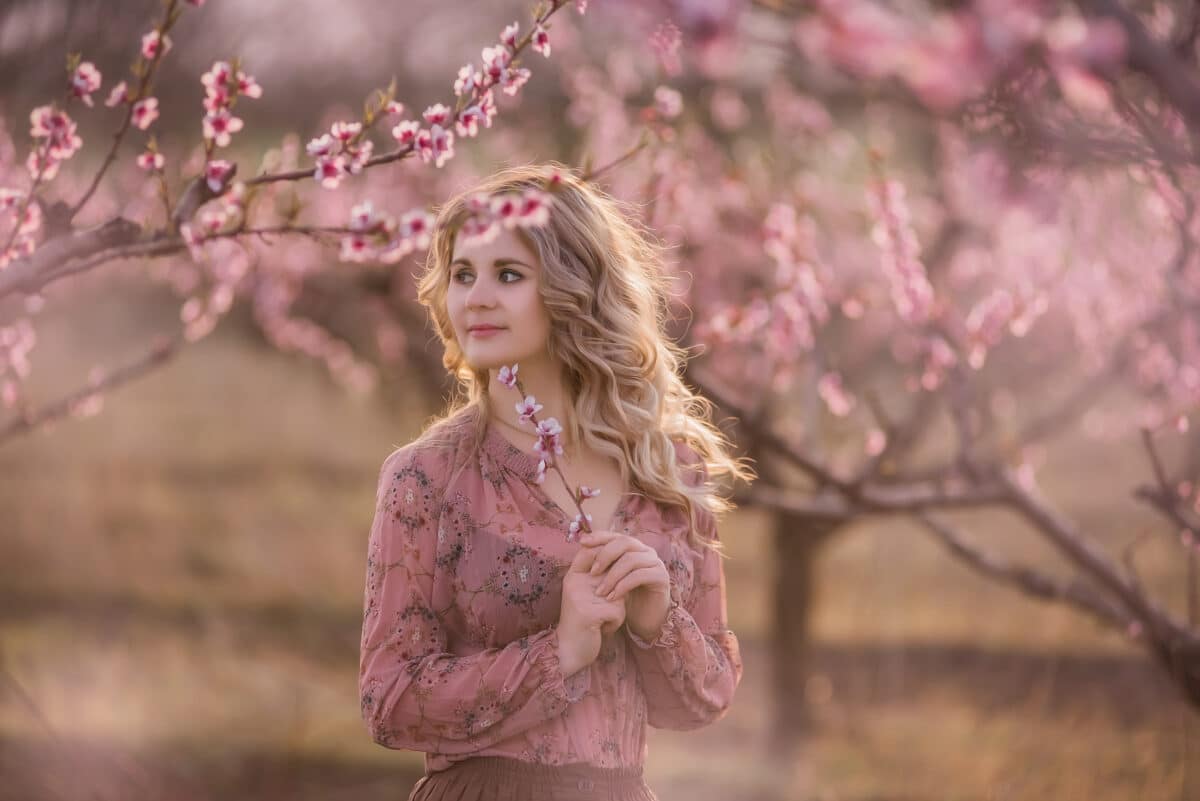 Tender fresh young girl with beautiful curly blonde hair, with a clean face, natural makeup, stands in blooming gardens, holds a twig with pink inflorescences. Mysterious, pensive, sensitive woman