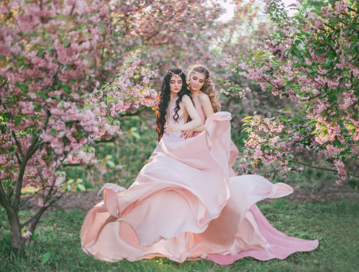 Two amazing elves walk in the fabulous cherry blossom garden. Princesses in luxurious, long, pink dresses that flutter in the wind. Two loving sisters embrace each other. Art photo, vanilla colors