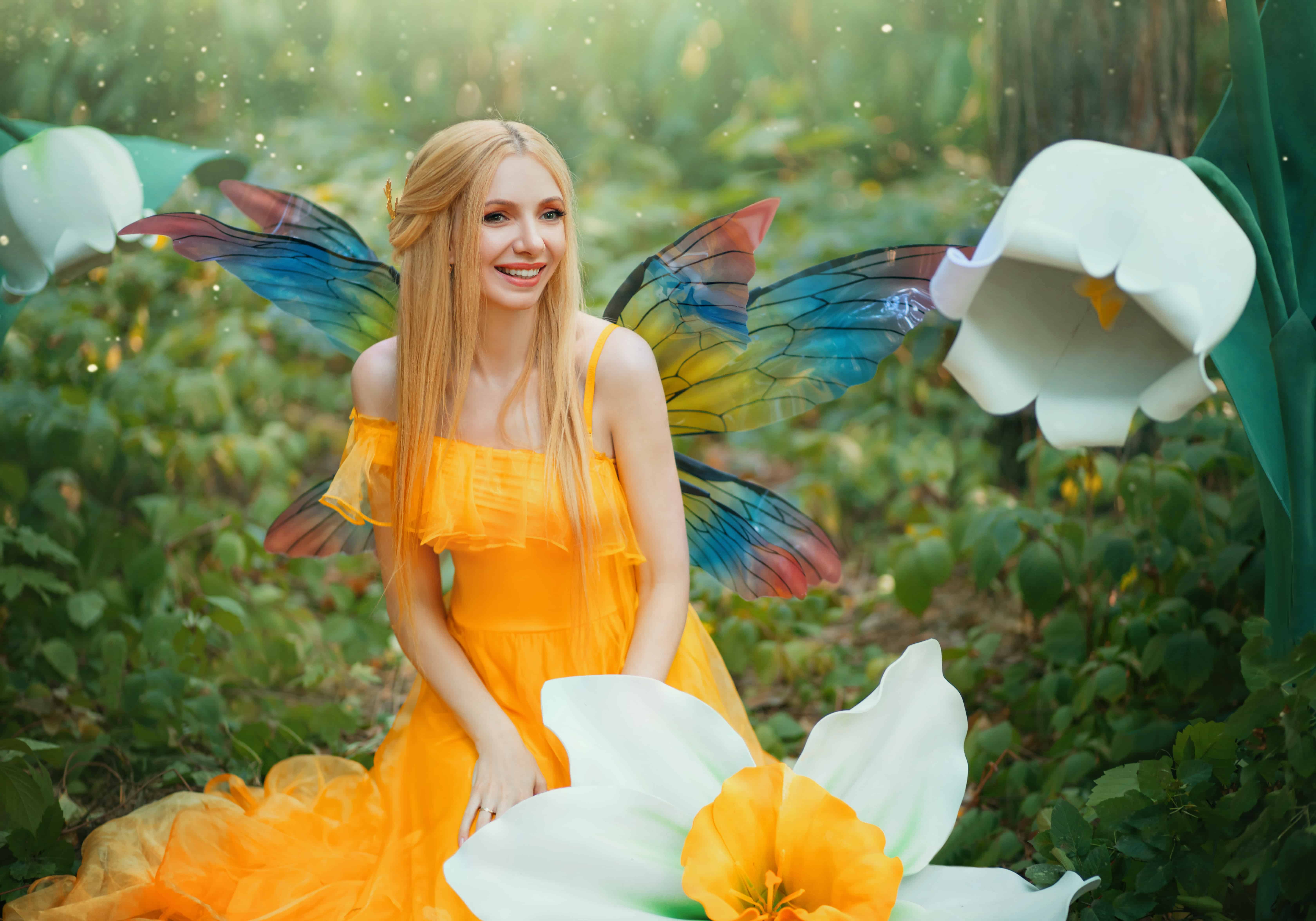 Beautiful happy woman fairy nymph sitting on forest. Magical fantasy wings costume pixie butterflies. Elf girl princess long yellow dress. blond hair smiling face. Summer nature tree, green grass