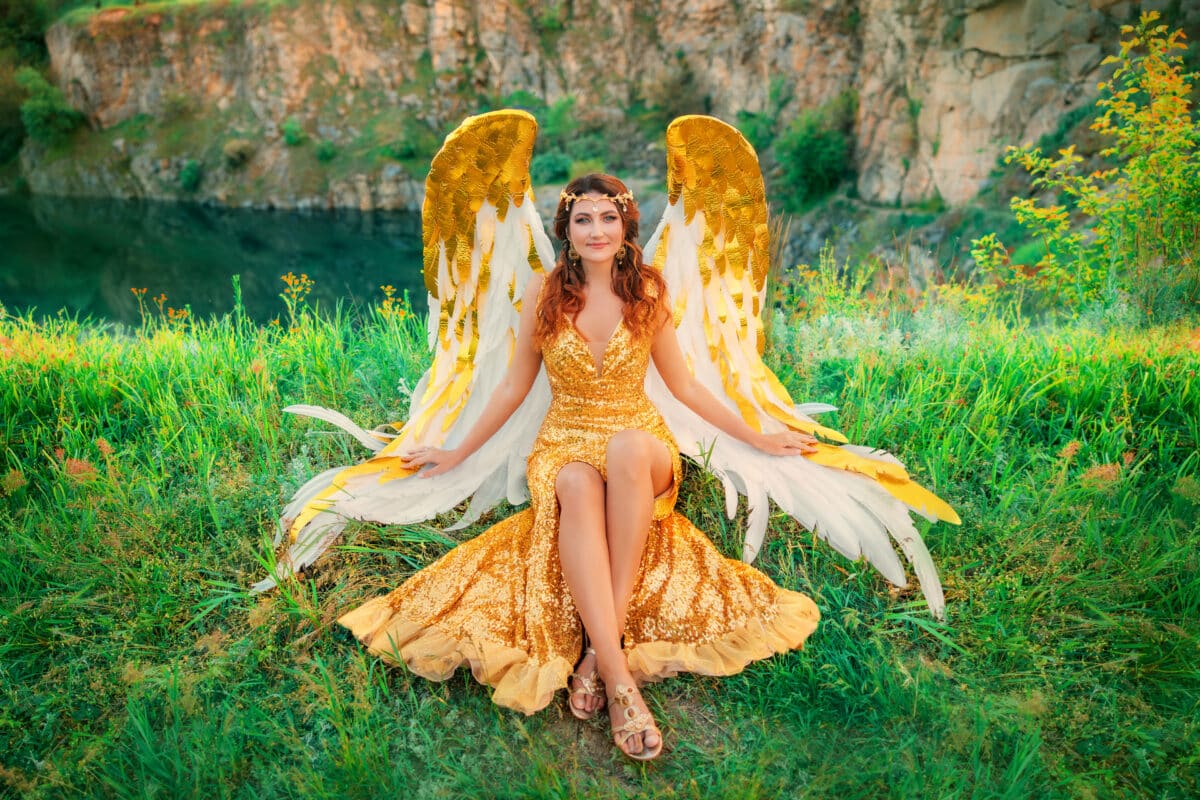 Art photo portrait happy redhead Fantasy woman goddess angel sits on river bank on green grass, magic divine light. golden white wings costume feather bird long gold dress. Fairy girl smiling face