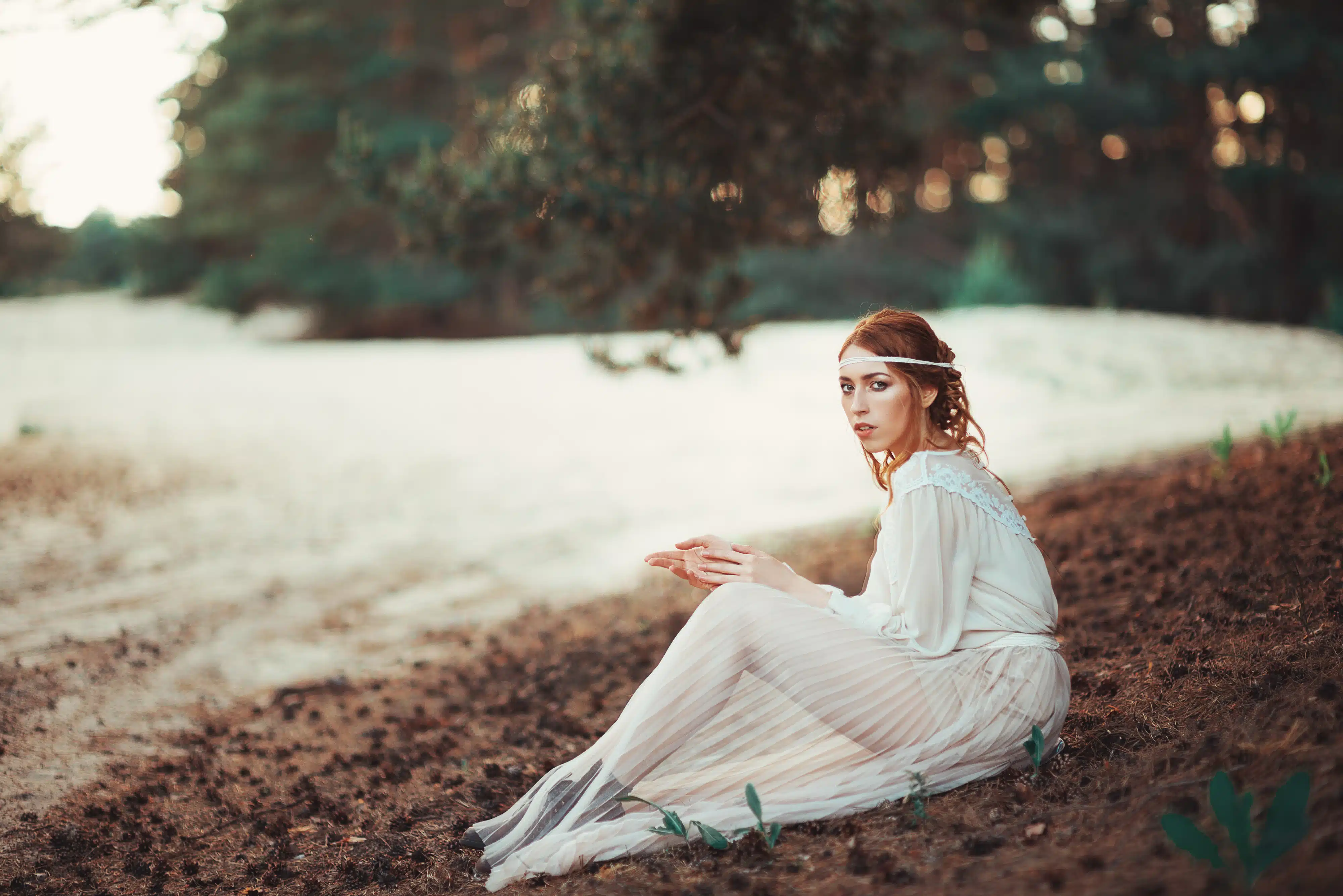Beautiful artistic photo portrait of a mysterious girl in white dress sitting on the shore of the lake near the forest