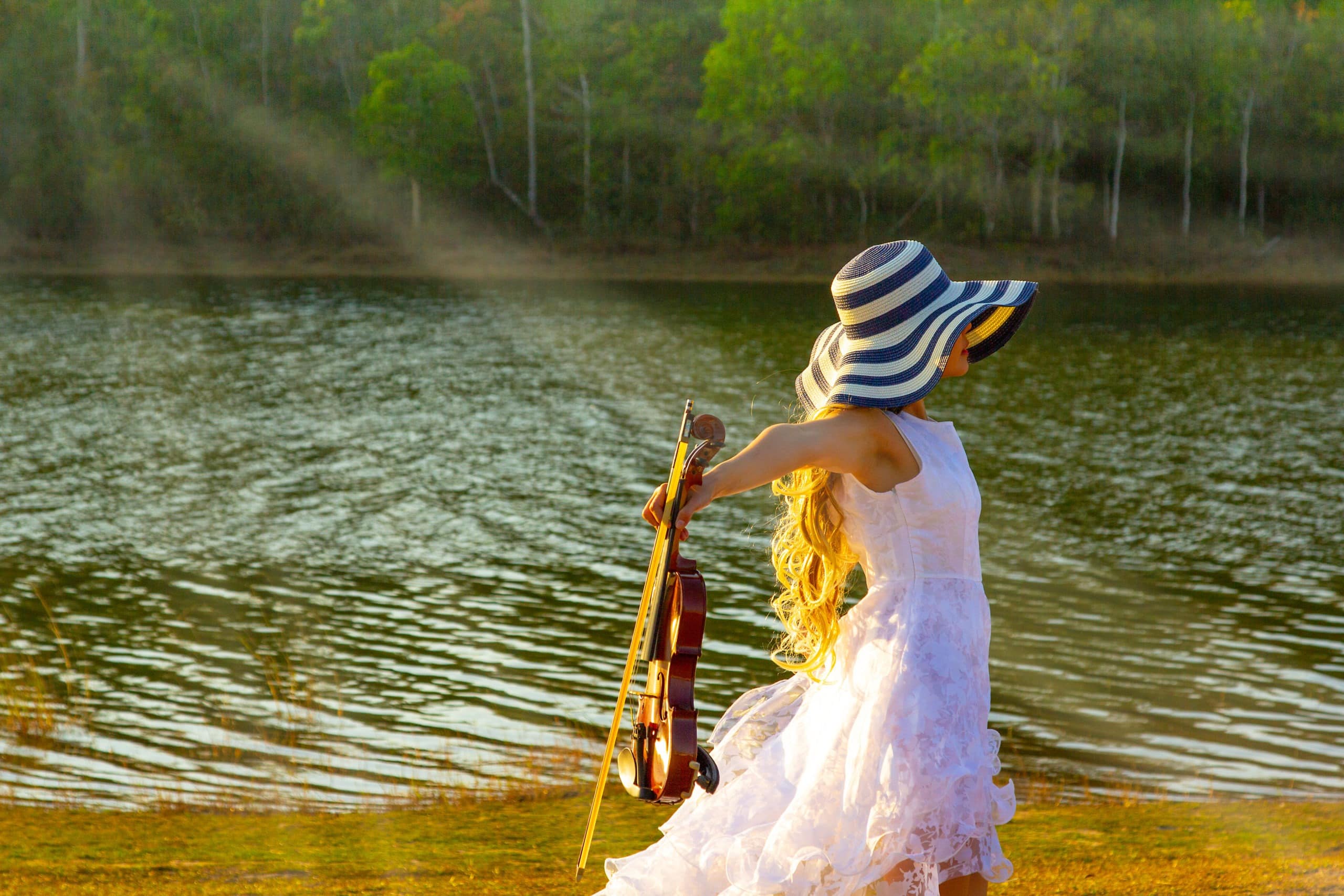 Woman in white dress and a hat holding the violin while standing by the lake.