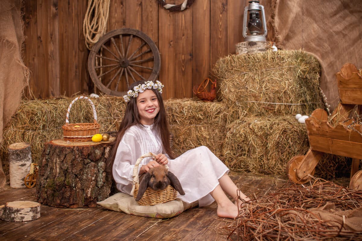 child in white dress and wreath of flowers on the head is sitting in the barn with a brown rabbit