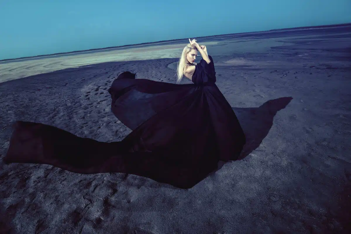 mysterious woman in black flowy dress standing on the sand by the lake