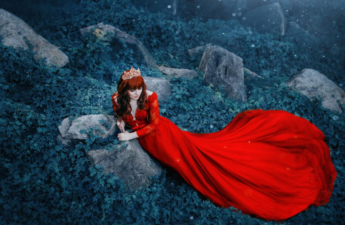 The Queen in a luxurious, expensive, red dress, with a long train lies on the thickets of ivy. Red-haired girl in a gold crown. The background is cold, juicy greens. Art photo