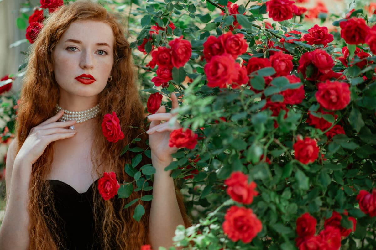 Beautiful redhead freckled woman with long curly hair, red lips makeup, wearing elegant pearl necklace, vintage dress, posing in rose garden. Copy, empty space for text