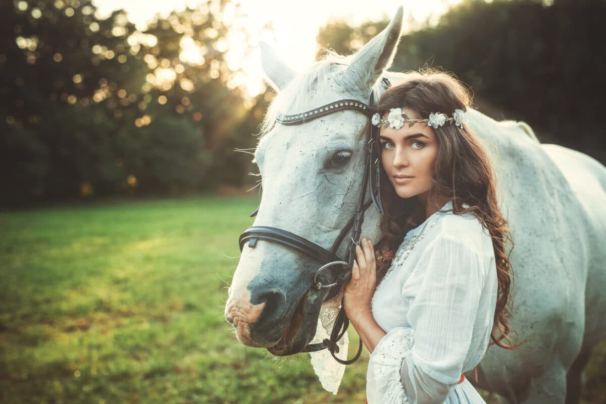 Beautiful young lady dressed in white with a flower head wreath standing beside her horse