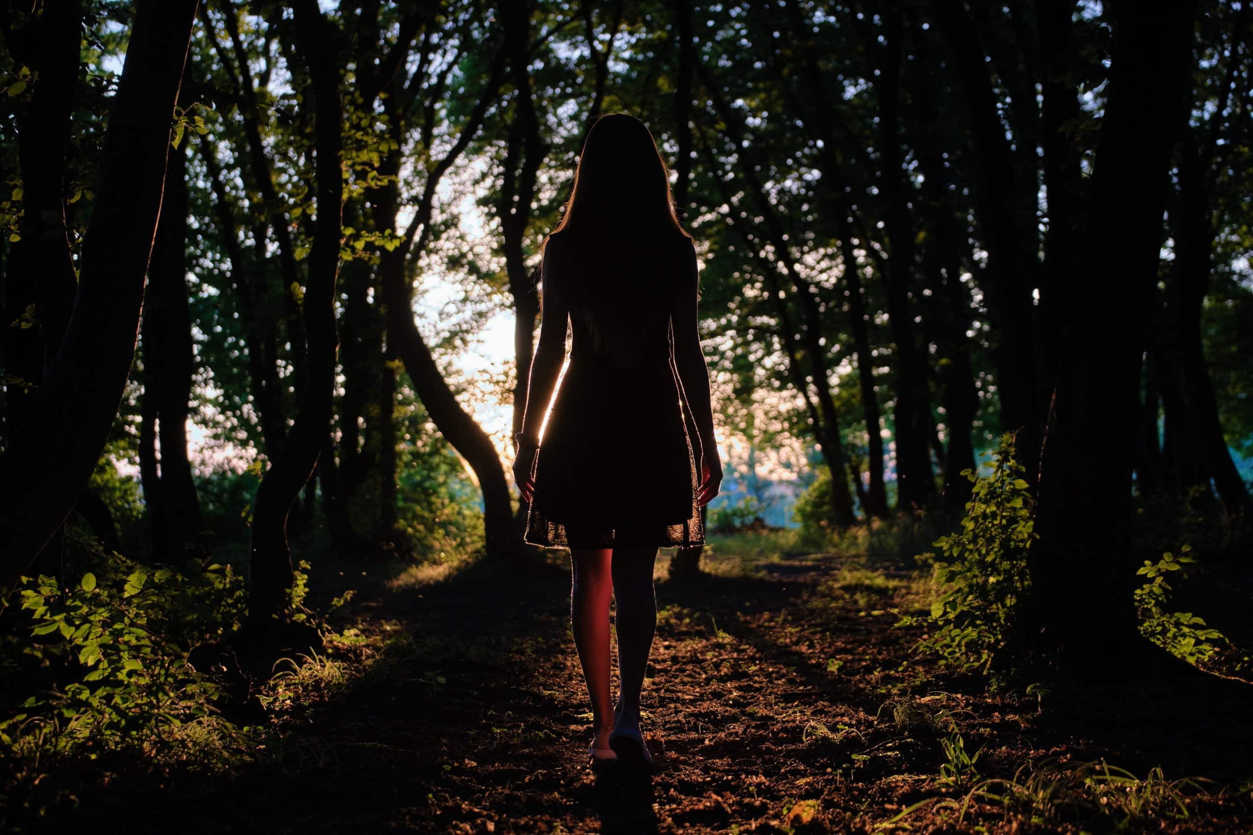 Young woman in dress walking alone through dark woods at bright sunset.