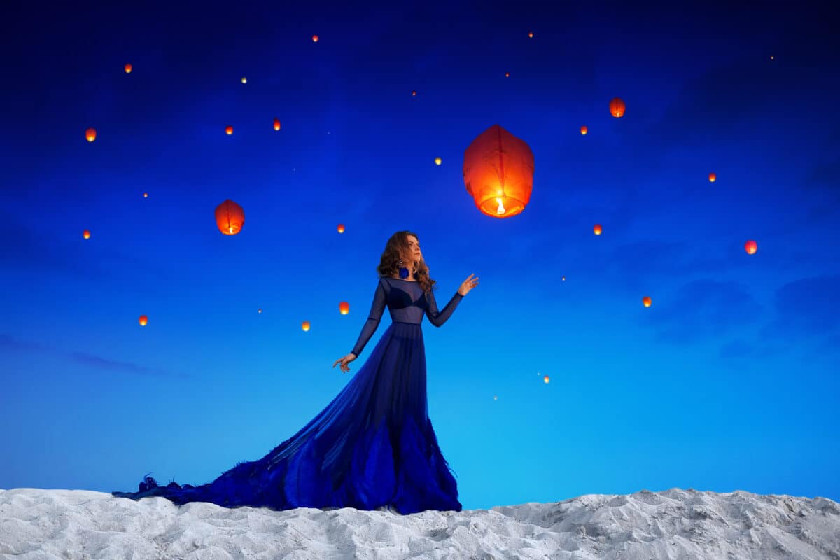 A beautiful girl in a blue long dress stands on white sand looking up at the floating lanterns in the sky