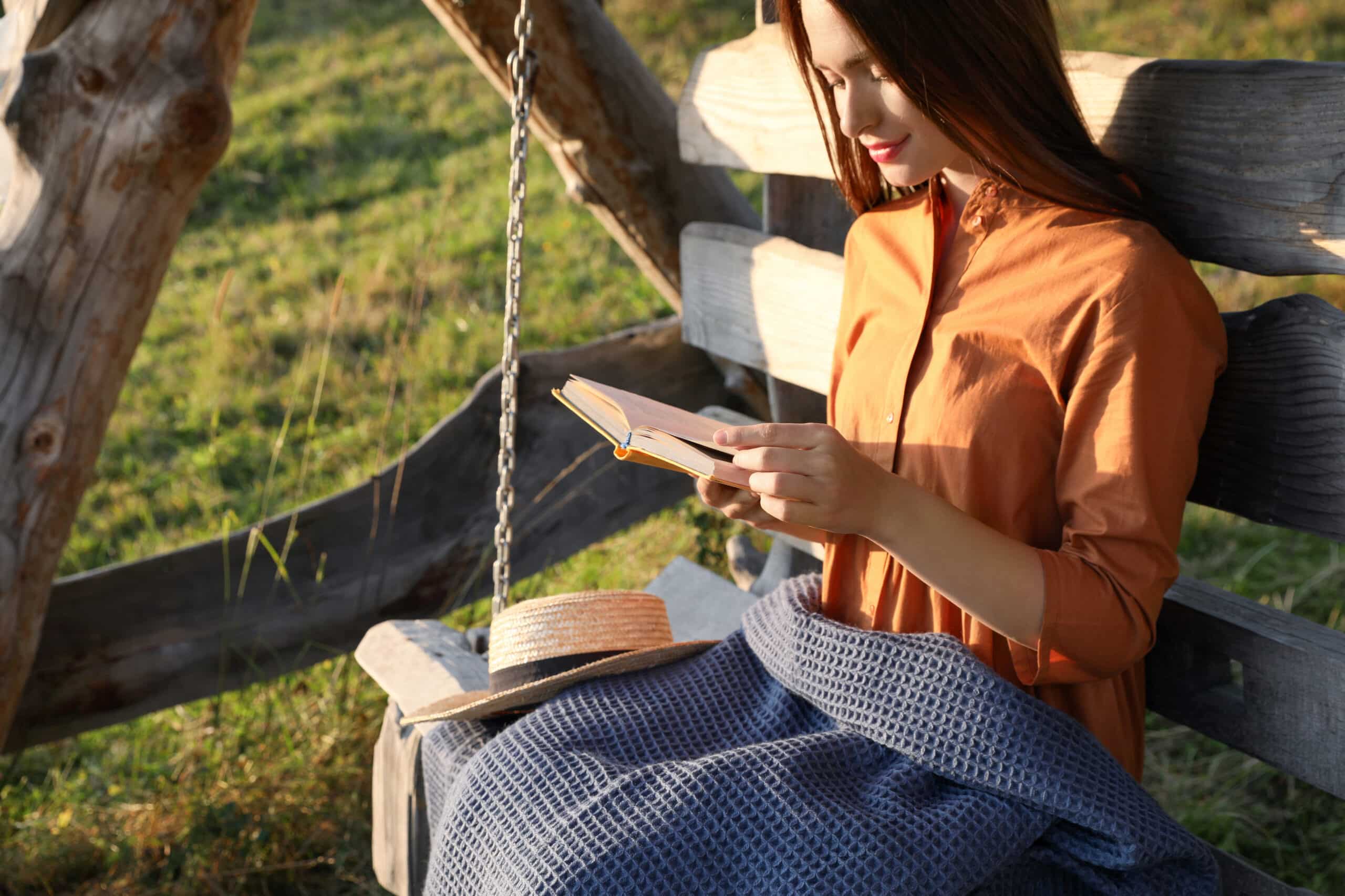 Beautiful young woman reading book on wooden swing outdoors