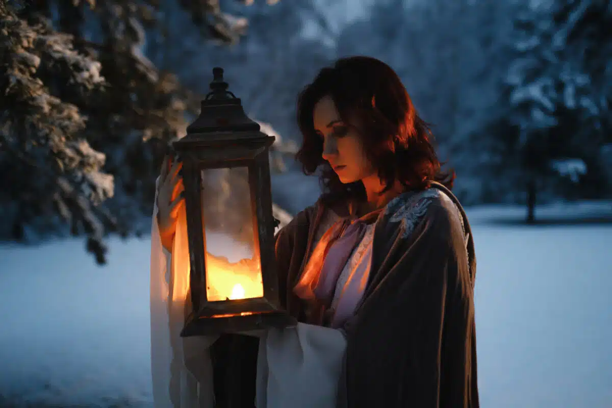 Beautiful fairytale shot of young woman looking at vintage lantern emitting warm light. Medieval fantasy concept