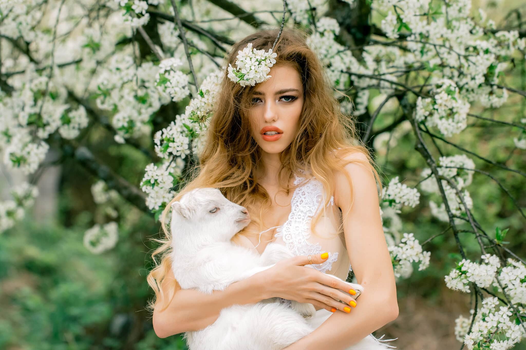 a young beautiful woman holding a small white lamb in spring blooming garden