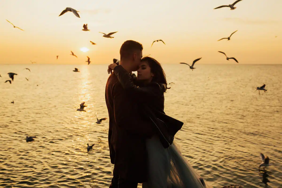 silhouette of a young couple in love hugging by the sea at sunset with seagulls hovering above the water