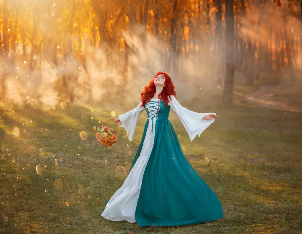 charming girl with bright red hair in national Bavarian green costume joyfully dances on green meadow near summer forest, magical sorceress collected fresh apples basket in rays of warm setting sun