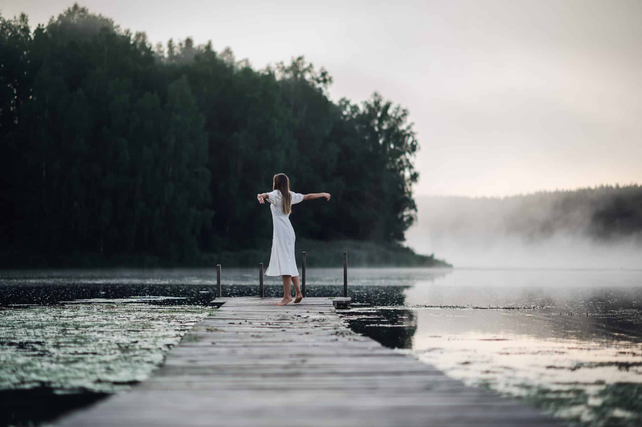 young woman in white dress standing alone on footbridge and staring at lake on a foggy morning