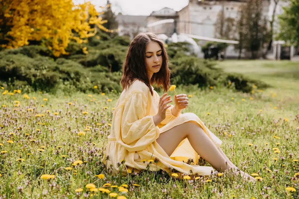 Young barefoot woman wearing a yellow muslin dress, sitting on the grass with yellow dandelions