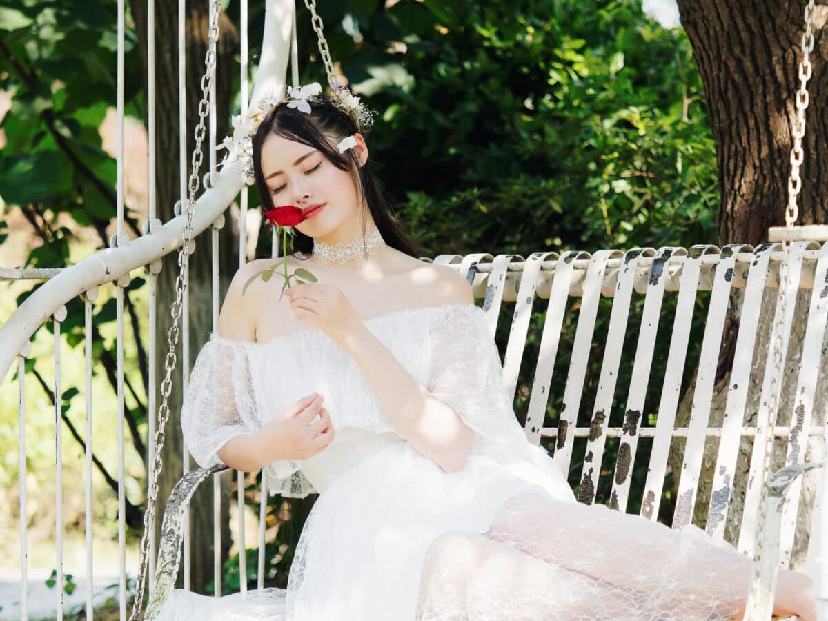 Beautiful fairy lady in white wedding dress and garlands of flowers, posing with red rose in hand, pretty Chinese girl sit in white chair in sunny day. Emotions, people, beauty and lifestyle concept.