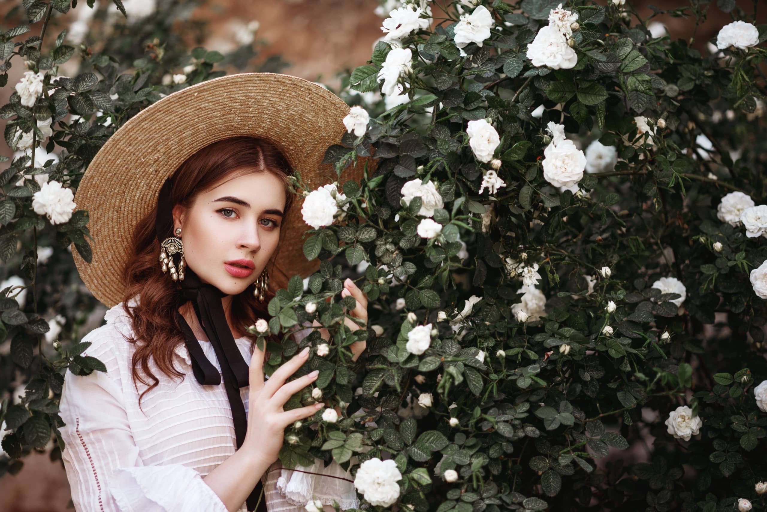 Young beautiful lady in white dress and big brown hat standing near a rose bush