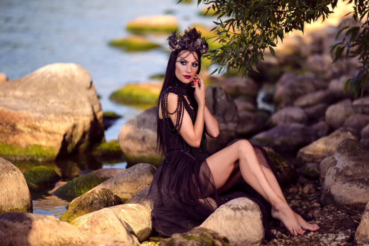a mysterious woman dressed in short black dress with a diadem is sitting on a rock by the river