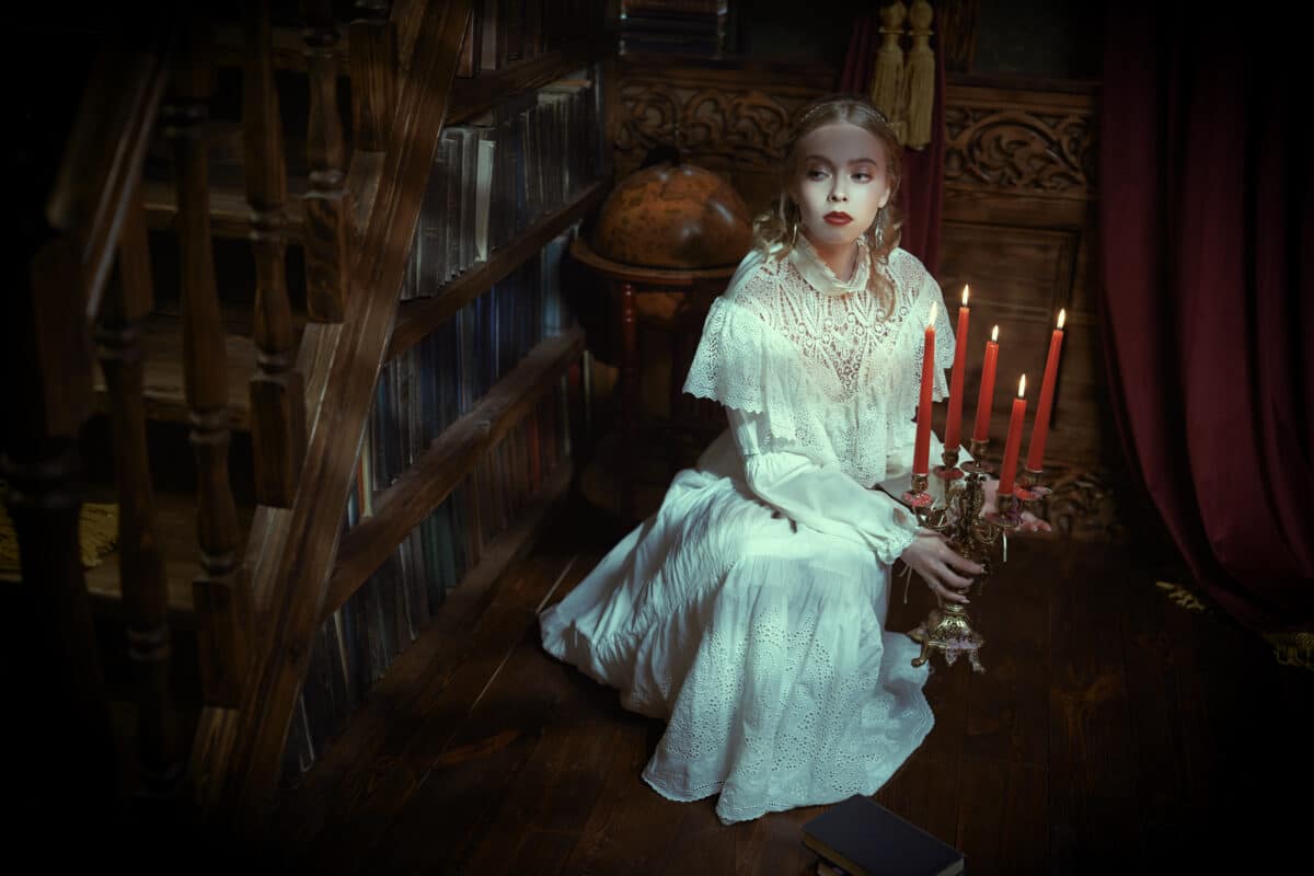 mystical lady aristocrat in a white lace dress sits in a dark vintage library by candlelight looking warily away