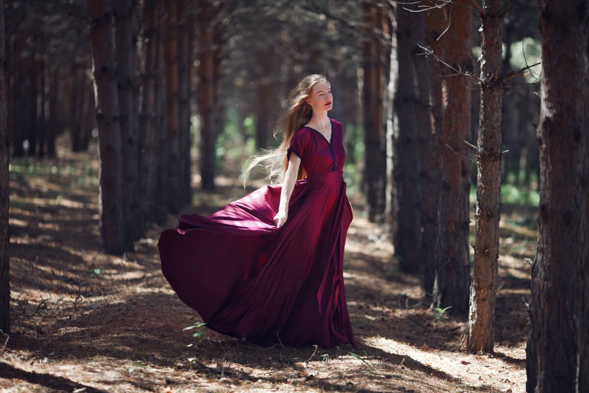 a tall, slender woman with long blond hair walks in the woods in a burgundy dress