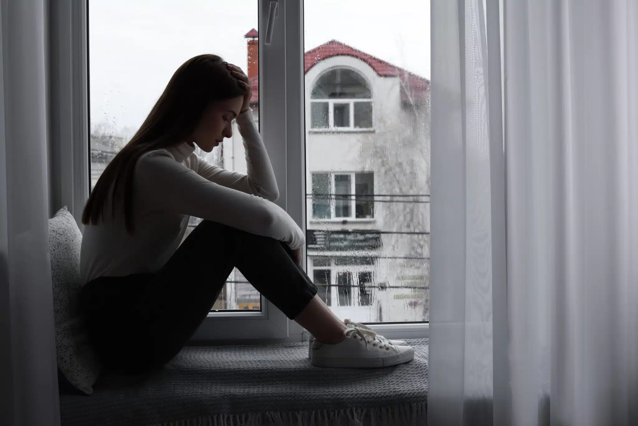 Unhappy young woman near window indoors, space for text.