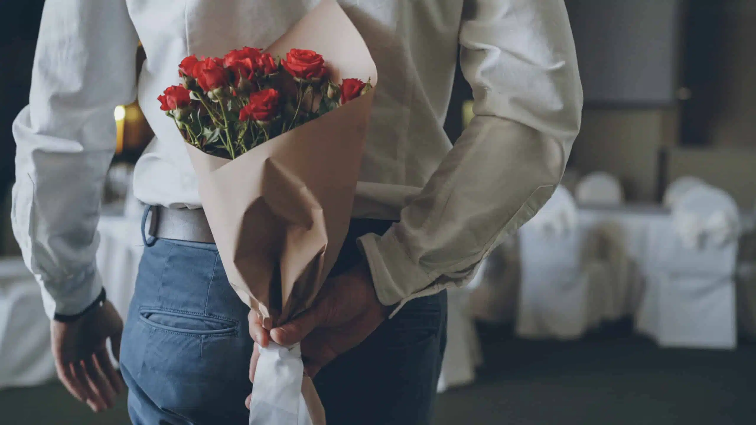 Loving man is hiding red roses behind his back bringing beautiful bouquet for his date in restaurant. Flowers, romantic relationship and dating concept.