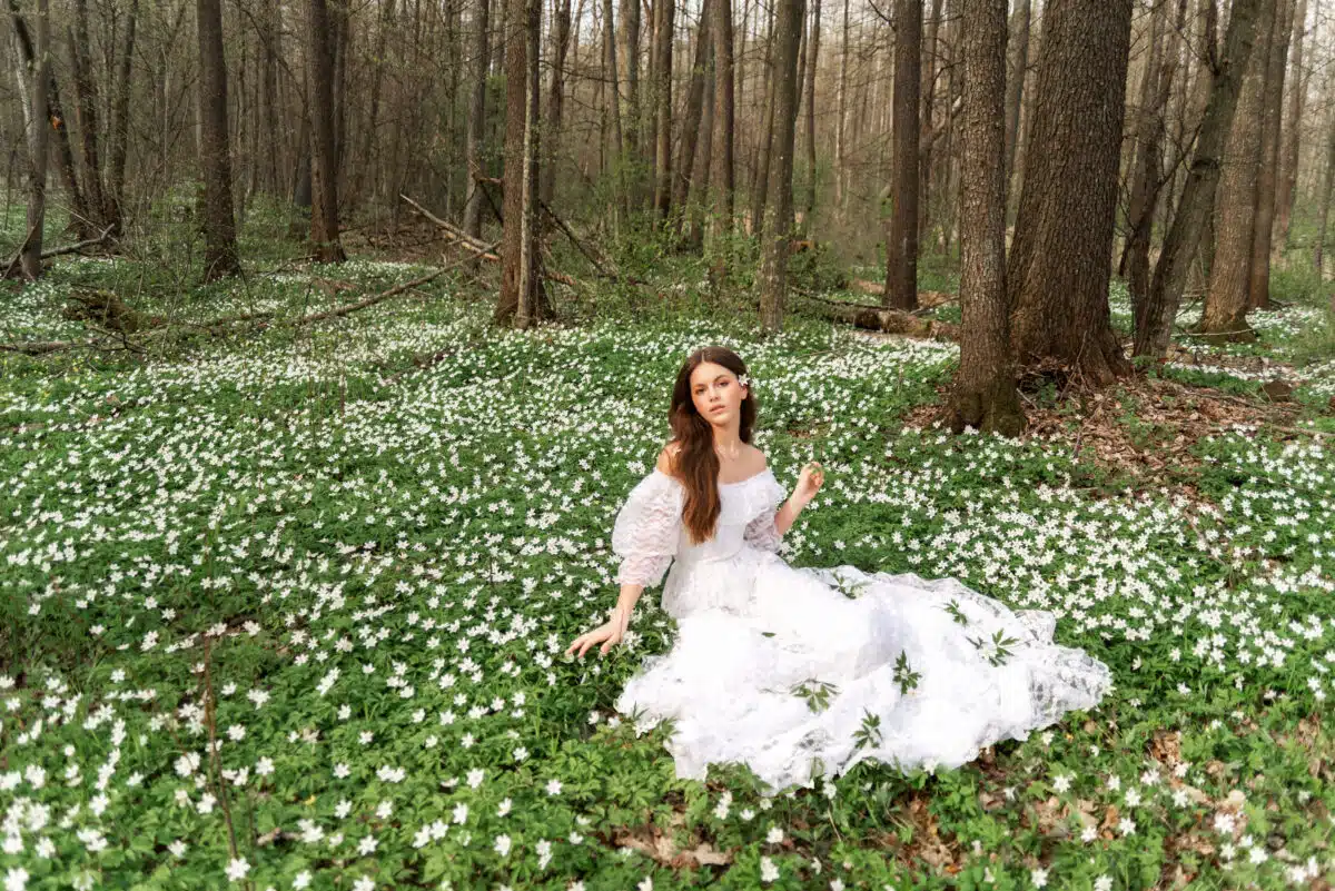 Girl in a white lace dress is sitting in a clearing with primroses. A young woman with long hair collects anemones in the spring forest.