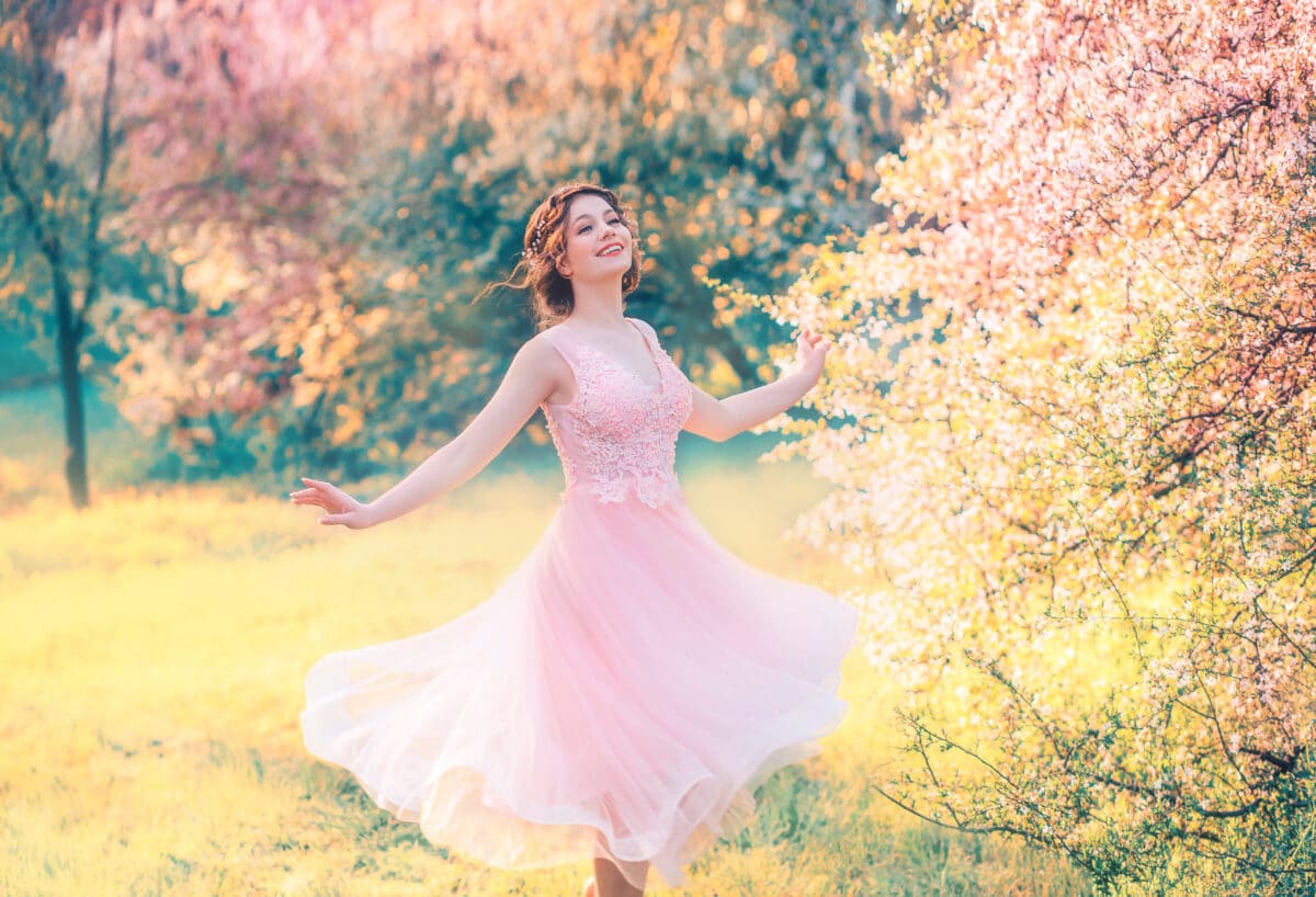 happy girl in short flying gentle pink dress laughs joyfully, doll princess whirls in bright yellow spring garden with flowering trees, positive emotions