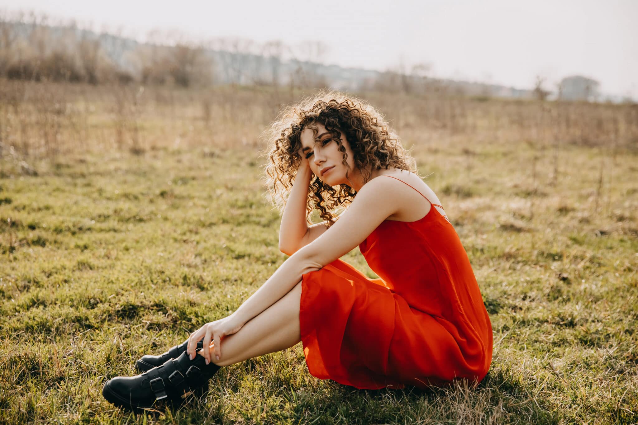 Young calm woman with curly hair, wearing a red summer dress outdoors, sitting in a field.