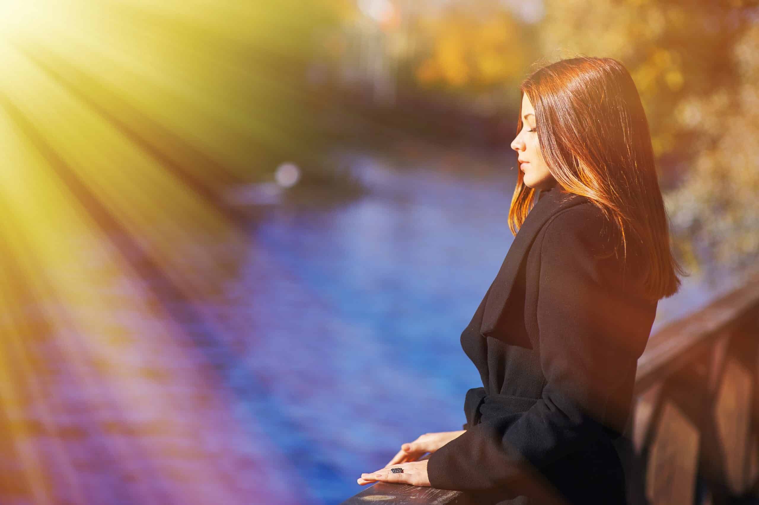 Portrait of young pensive female with a gorgeous shiny hair stands the embankment of river with closed eyes in sunset on blurred background autumn Park, closeup.